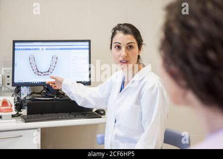Junge weibliche Stomatologin im medizinischen Kleid sitzt auf Stuhl und Zeigt die Zähne des Patienten am Computer in der Zahnmedizin Stockfoto
