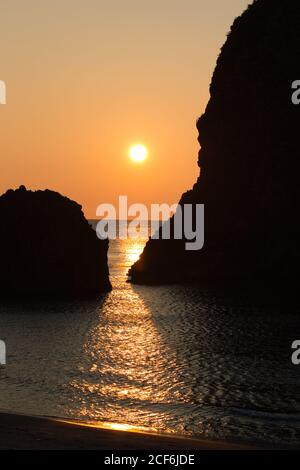 Sonnenlicht scheint zwischen zwei großen Felsen im Meer und Reflektiert in den ruhigen Gewässern, während die Sonne untergeht Stockfoto