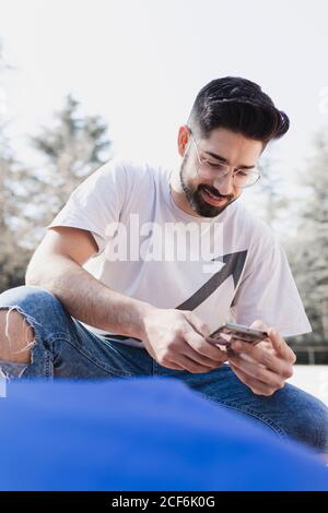 Von unten junger bärtiger Kerl in Brillen und Freizeitkleidung Surfen auf dem Handy im Park bei sonnigem Wetter Stockfoto