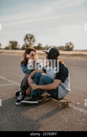 Trendiges Teenager-Paar auf Schlittschuhe auf leerer abgelegener Straße sitzen Und sich gegenseitig anschauend reden Stockfoto
