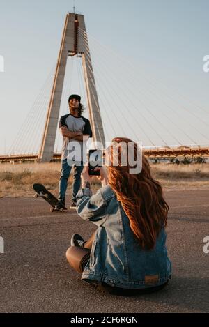 Langhaarige stilvolle Teenager weiblich Schießen cool Hipster Kerl mit Skateboard gegen moderne Brücke im Grünen Stockfoto