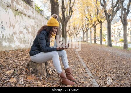 Afroamerikanische Frau in gelbem Hut und warmer Jacke mit Smartphone auf der Straße mit Herbstblättern im Park sitzen Stockfoto