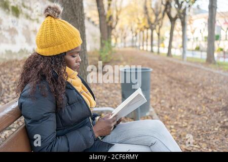 Seitenansicht einer nachdenklichen, stilvollen afroamerikanischen Frau in gelbem Hut und warmer Jacke bequemes Sitzen auf Holzbank und Lesen von Büchern im Herbstpark Stockfoto