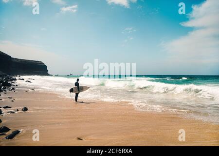 Ganzkörper anonymer Mann mit Surfbrett steht am Sandstrand in der Nähe winkenden Meerwassers an sonnigen Tag auf Fuerteventura Insel, Spanien Stockfoto