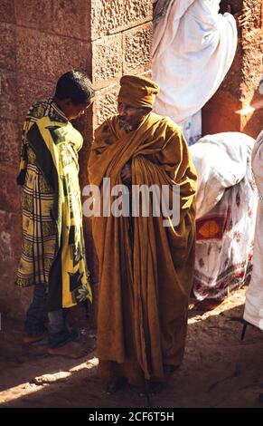 Lalibela, Äthiopien - 04. November 2018: Älterer ethnischer Mann im langen Gewand, der auf Felsen einer alten monolithischen Kirche mit Gebeten steht, Äthiopien Stockfoto