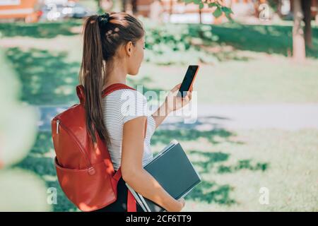 Zurück zum Schulsemester online per Telefon-App. Universitätsstudentin, die ihr Handy benutzt, um ihren Unterricht zu sehen, während sie mit Rucksack auf dem Campus spazieren geht Stockfoto