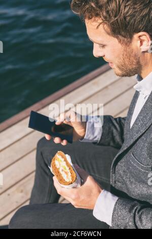 Professioneller Mann in der Mittagspause von der Arbeit mit Handy Essen Sandwich draußen am Kanal Wasser. Kaukasischer junger Erwachsener mit Smartphone-Handy Stockfoto