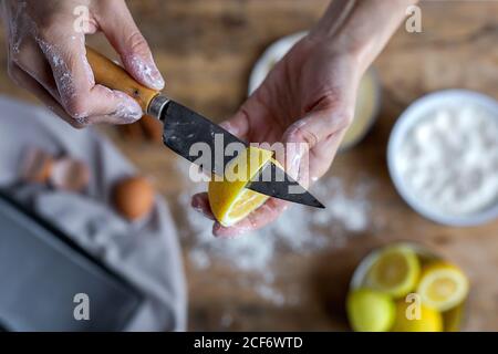 Von oben Ernteguthand einer nicht erkennbaren Frau, die mit Mehl bedeckt ist, das Zitrone schält und der Kamera eine frische, halbgeschnittene Zitrone mit einem Messer zeigt Stockfoto