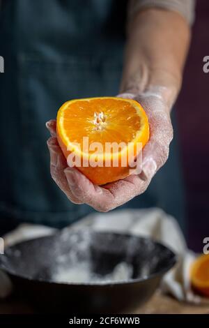 Die Hand einer nicht erkennbaren Frau, die mit Mehl bedeckt ist, hält und der Kamera eine frische, halb geschnittene Orange über der Schüssel zeigt, während der Teig am Tisch zubereitet wird Stockfoto
