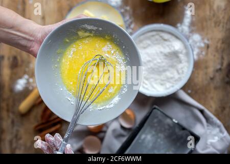 Von oben Ansicht der Ernte anonyme Frau schlagen Eier in schwarzer Schüssel auf Holztisch mit Zitrone, Mehl, Butter und Zimt Sticks Zutaten für Kuchen Stockfoto