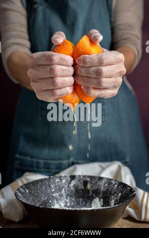 Ernte Hand von unkenntlich Frau in Schürze quetschen frisch saftig geschnitten Orange über Schüssel während der Zubereitung von Teig am Tisch Stockfoto