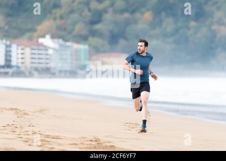 Bärtiger männlicher Athlet im aktiven Tragen beim Laufen bei leerem Sand Strand mit grünen Bergen auf verschwommenem Hintergrund Stockfoto