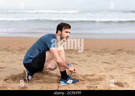 Seitenansicht des bärtigen sportlichen Mannes im aktiven Sitzen Hocken und binden Schnürsenkel auf sandigen leeren Meer und suchen Weg Stockfoto