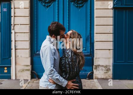 Seitenansicht der glücklich junge Paar in lässiger Kleidung Umarmung Und küssen, während sie gegen gealtertes Steingebäude mit Blau stehen Türen auf der City Street Stockfoto