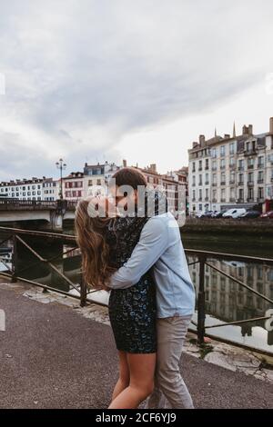 Seitenansicht der glücklichen jungen liebevollen Paar umarmen und küssen Auf einem Steindamm in der Nähe des Flusses mit alten Gebäuden Im Hintergrund in Bayonne Stadt in Frankreich Stockfoto