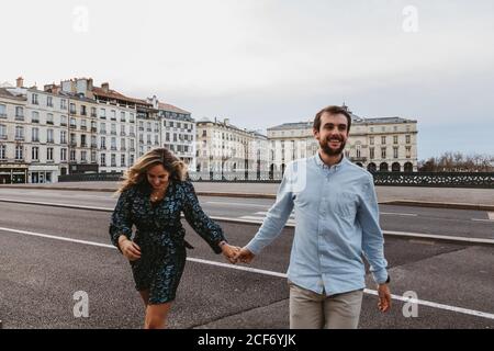 Glücklich junge romantische Paar in stilvollen Kleidern lachen und halten Hände beim Überqueren Brücke mit historischen Gebäuden im Hintergrund während Stadtrundfahrt in Bayonne in Frankreich Stockfoto