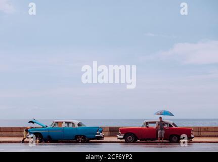 Havanna, Kuba - 14. DEZEMBER 2019: Ein Mann mit Regenschirm steht neben einem roten Retro-Auto, während ein anderer Mann an einem regnerischen Tag am Meer mit bewölktem Himmel im Hintergrund ein altes blaues Auto repariert Stockfoto