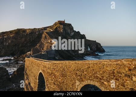 Leere Straße entlang Steinbrücke und Kamm von felsigen Hügel zu einsamen Landhaus auf Insel Gaztelugatxe umgeben von Ruhiges Meerwasser mit weißen Schaumwellen während des Sonnenuntergangs Stockfoto