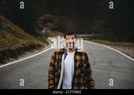 Bärtige schwarzhaarige fröhliche Mann in lässigen tragen lächelnd und Blick auf die Kamera, während Sie im Herbst auf einer leeren Straße stehen Auf dem Land Stockfoto