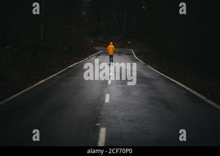 Rückansicht des anonymen Mannes in gelber Jacke, der auf geht Leere Asphaltstraße zwischen grünem Wald Stockfoto