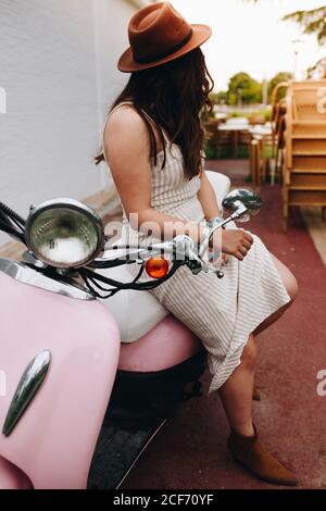 Seitenansicht einer nicht erkennbaren modischen Frau, die auf einem rosa Motorrad auf der Straße sitzt Stockfoto
