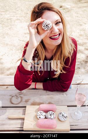 Junge attraktive lächelnde Frau, die durch das Donut-Loch schaut, während sie am Picknicktisch sitzt Stockfoto