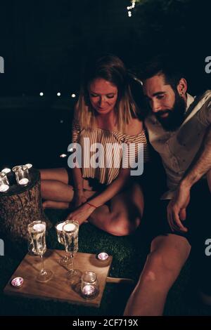 Junger Mann und junge Frau sitzen auf dem Rasen in der Nähe glühender Kerzen während der Nacht im dunklen Nachtgarten Stockfoto