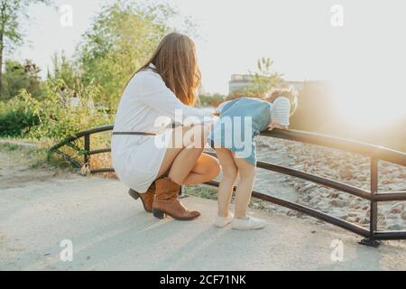 Seitenansicht einer jungen Frau und eines kleinen Mädchens, das sich über das Geländer beugte und den schnellen Wasserstrahl an einem sonnigen Tag im Park ansah Stockfoto