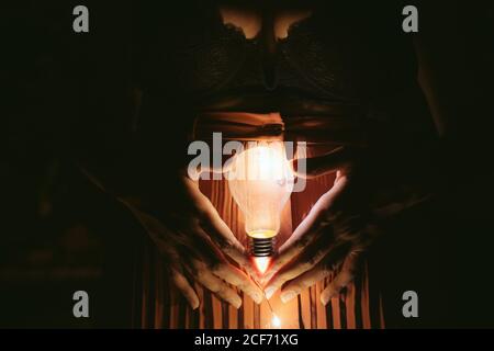 Anonyme Frau in BH und Rock hält brennende Glühbirne mit Händen in der Dunkelheit Stockfoto