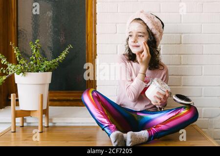 Positive kleine Mädchen in legerer Kleidung und rosa Stirnband sitzen Auf dem Tisch mit Topfpflanze und Anwendung Gesichtsmaske aus Glas vor Hintergrund der weißen Ziegelwand mit Holz Angezeigt Stockfoto