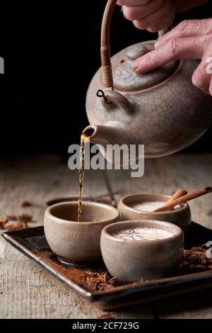 Crop Person mit Teekanne Ausgießen Masala chai in Keramikschalen Auf Tablett mit Sternanis und Zimtstangen gelegt Stockfoto
