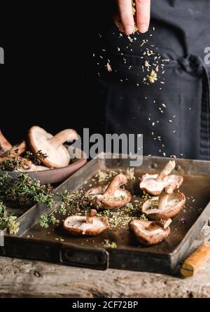 Crop Person bestreuen mit Kräutern frischen braunen Pilzen Shiitake auf Metalltablett am rustikalen Holztisch Stockfoto