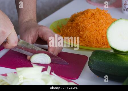 Ein Mann zerkleinert eine Zwiebel. In der Nähe Gemüse zum Kochen Squash Kaviar. Zucchini, Karotten, Zwiebeln und Tomaten auf der Tischoberfläche. Stockfoto