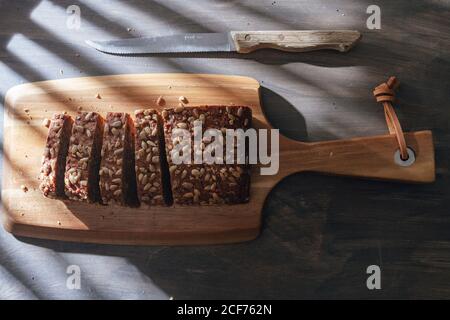 Draufsicht auf geschnittenes Brot aus Vollkornbrot auf Holz Schneidebrett und Küchensäge Messer auf Holztisch platziert Mit Sonnenlicht und gestreiften Schatten Stockfoto