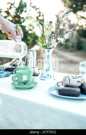 Crop Dame Gießen Milch aus transparenten Krug zu grünen Tasse Beim Kaffee am Tisch mit Wildblumen in Vase und Frische Donuts auf dem Teller ruhen in der Natur Stockfoto