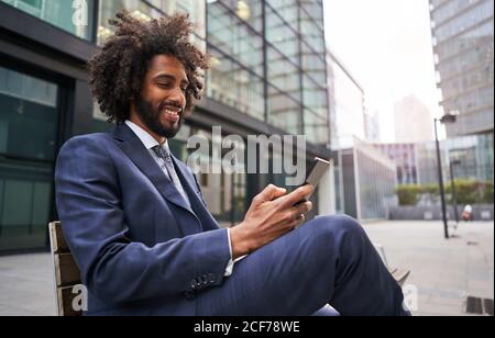 Glücklicher schwarzer Angestellter mit afro Haaren sitzt auf Bank und Lächelnd beim Surfen in sozialen Medien mit dem Smartphone Stockfoto