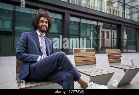 Optimistischer und fröhlicher afroamerikanischer Unternehmer mit Bart sitzend Bank mit gekreuzten Beinen und wegschauen Stockfoto