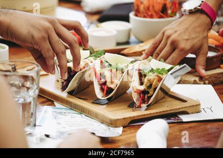 Crop anonyme Menschen essen köstliche traditionelle mexikanische Tacos serviert auf Holztablett auf dem Tisch mit verschiedenen Speisen und Getränken Stockfoto