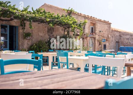 Holztische in Komposition mit weißen und blauen Stühlen Café-Terrasse gegenüber dem Äußeren der alten Gebäude dekoriert Topfblumen Und Klettern grünen Reben in sonnigen Tag Stockfoto