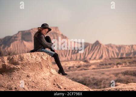 Seitenansicht einer jungen Frau in trendigem Outfit, die Knie umarmt und auf einer trockenen Klippe auf einem verschwommenen Hintergrund der Wüste von Bardenas Reales in Navarra, Spanien, sitzt Stockfoto