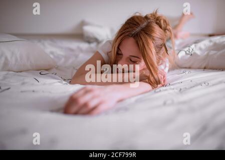 Junge zufriedene Weibchen mit Bleistift in roten Haaren, die Ruhe haben Nach dem Aufwachen am Wochenende Morgen und nach Hause liegen auf dem Bauch Auf gemütlichem Bett mit weißer Bettwäsche und lächelnd mit geschlossen Augen Stockfoto