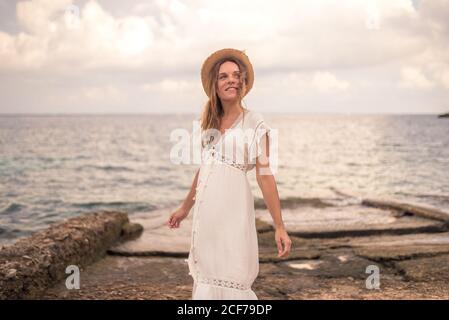 Unbeschwerte weibliche Reisende in Sommerkleid und Hut stehen auf Steinige Küste vor dem Hintergrund der erstaunlichen Seestang während des Urlaubs Stockfoto