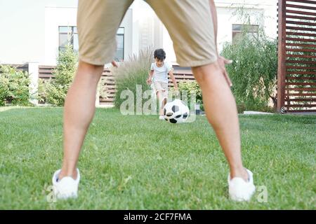 Nicht erkennbarer Mann spielt Fußball mit aktiven Jungen auf grünem Gras im Park, horizontal zwischen den Beinen erschossen Stockfoto
