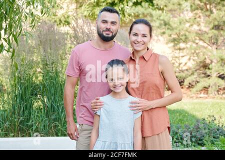 Familienportrait von Vater, Mutter und Tochter in lässigen Sommer-Outfits stehen zusammen im Park Blick auf Kamera lächelnd Stockfoto
