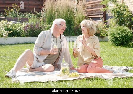 Der ältere Mann und seine liebenswerte Frau sitzen auf dem Rasen Hinterhof trinkt frische Limonade und plaudert Stockfoto