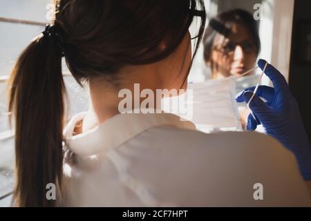 Rückansicht des anonymen weiblichen Spiegels im Spiegel in der Nähe Fenster und Anziehen einer schützenden medizinischen Maske während des Coronavirus-Ausbruchs Stockfoto