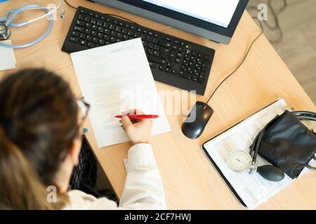 Seitenansicht der jungen Ärztin in Uniform beim Betrachen Papier und Tisch mit Tonometer im Zimmer Stockfoto