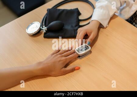 Von oben die Hände des weiblichen Arztes und des Patienten mit ernten Elektrischer Tonometer am Finger am Tisch in der Nähe des Pulsometers Stockfoto