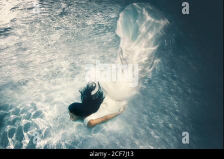 Schöne langhaarige Brünette in langen weißen Hochzeitskleid mit Schleier schwimmend unter Wasser mit ausgestreckten Armen Stockfoto