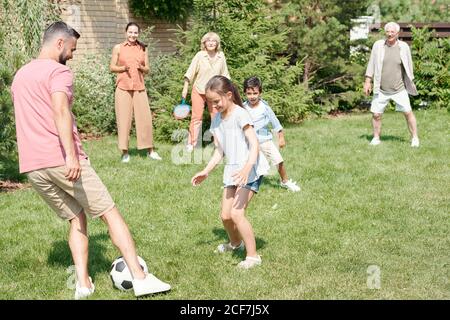 Granpa, Vater und zwei Kinder spielen Fußball auf Rasen im Park, Oma und Mutter beobachten sie Stockfoto